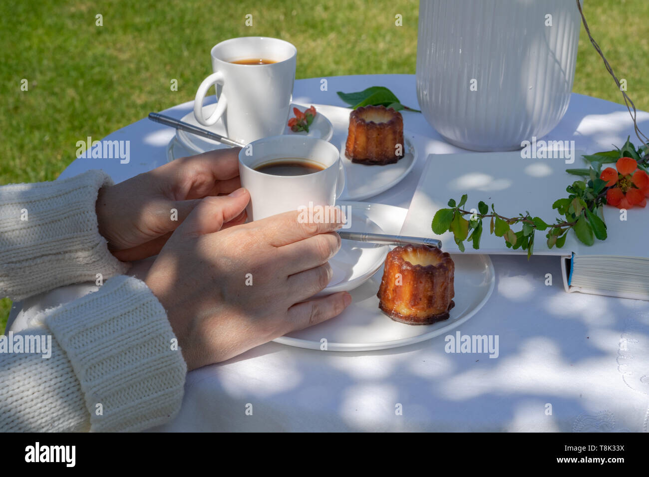 Momento di relax in una giornata di sole in giardino. Godendo di un caffè con caneles. Disposizione con una donna con le mani in mano e un bianco servito tabella. Foto Stock