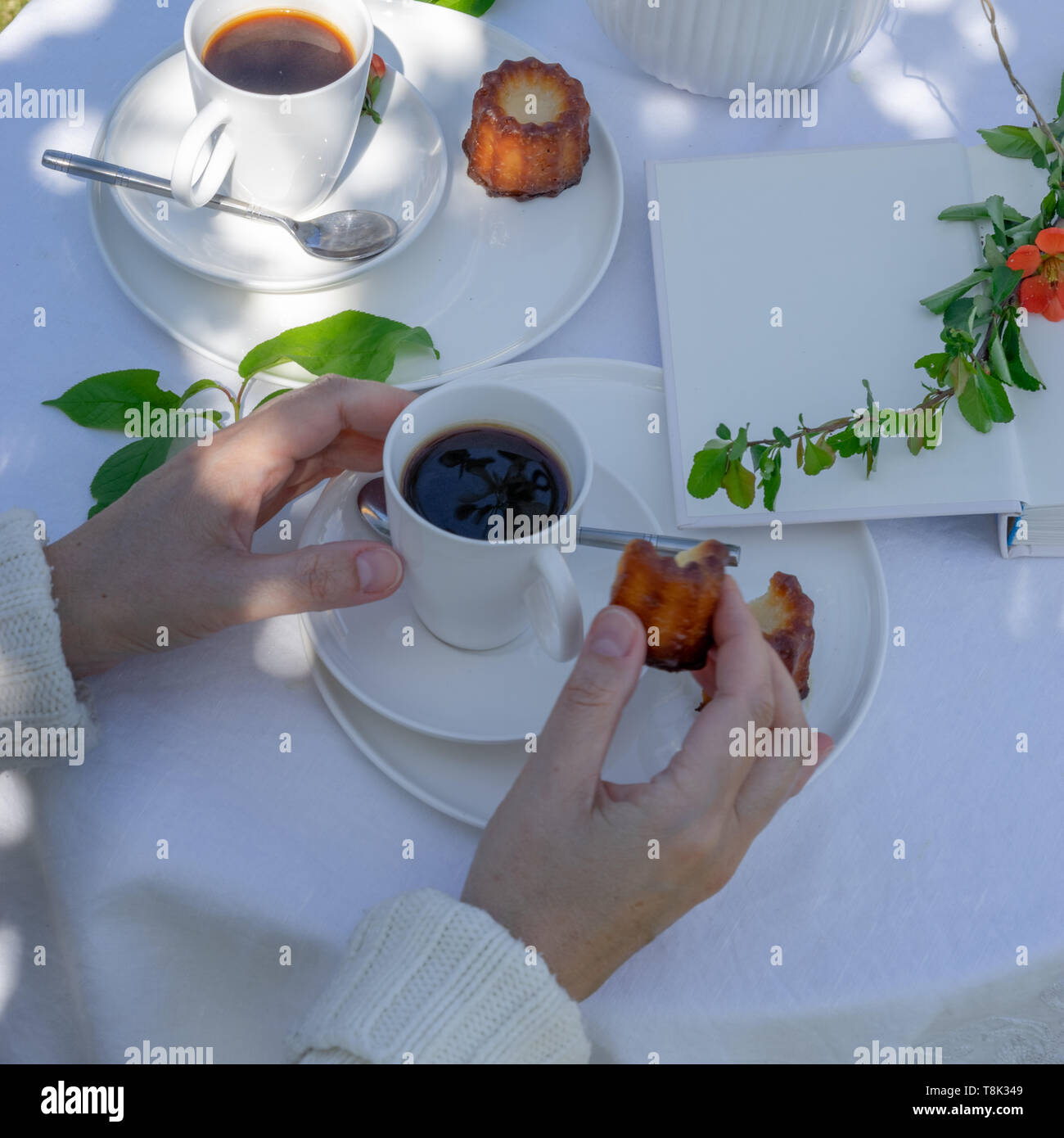 Bere un caffè di tempo in giardino. Tabella servita con caffè, canele, fiori e prenota in giardino. Disposizione con una donna con le mani in mano. Foto Stock