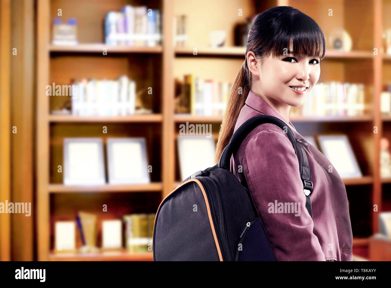 Asian student donna con zaino in piedi e guardare indietro nella biblioteca universitaria. Si torna a scuola concept Foto Stock