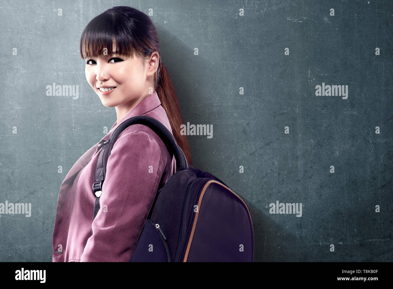 Asian student donna con zaino in piedi e guardare indietro con blackboard sfondo. Si torna a scuola concept Foto Stock