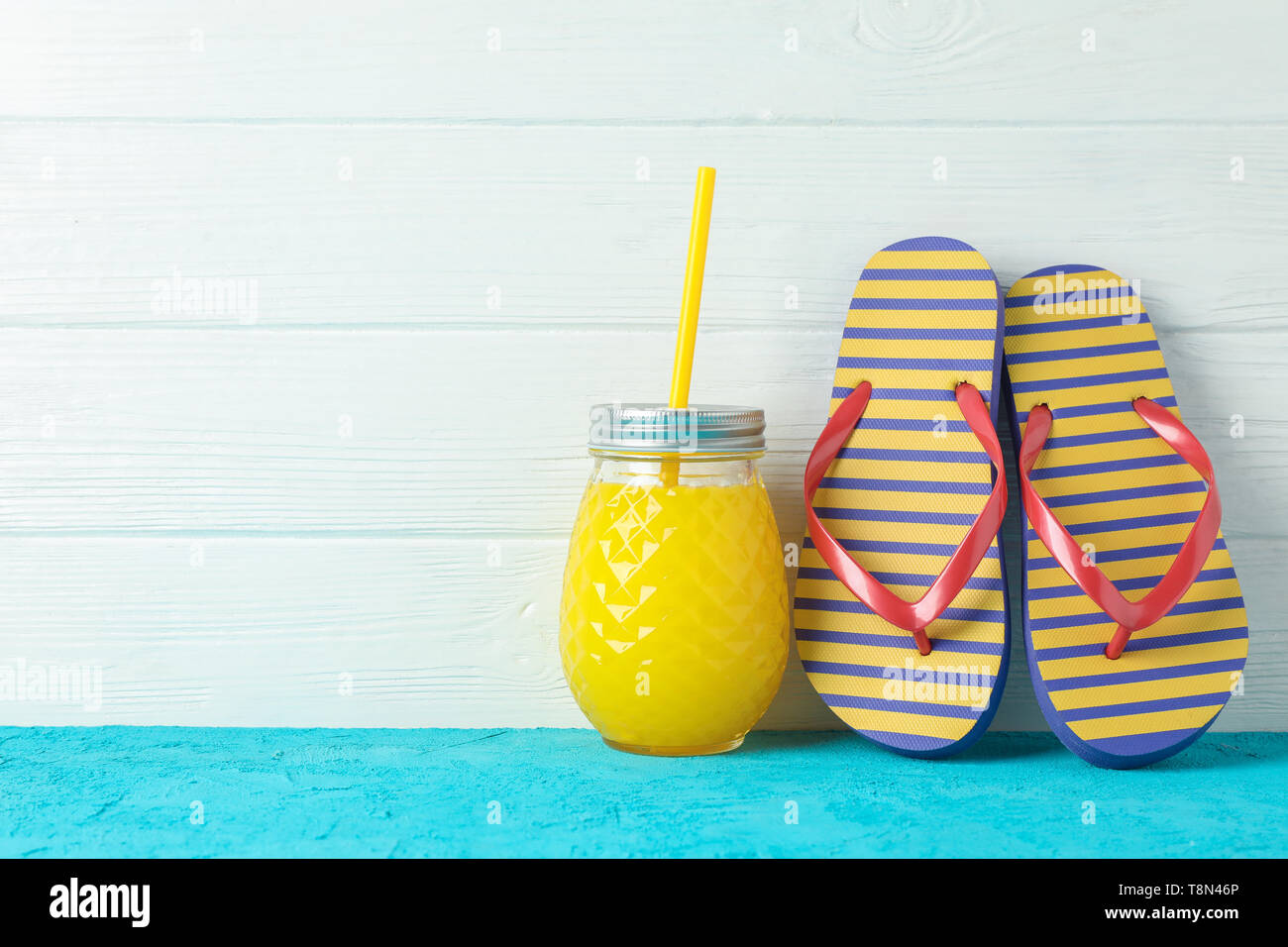 Il flip flop e vaso di vetro con del succo di frutta fresco sulla tabella di colore bianco contro lo sfondo di legno, lo spazio per il testo. La vacanza estiva sfondo Foto Stock