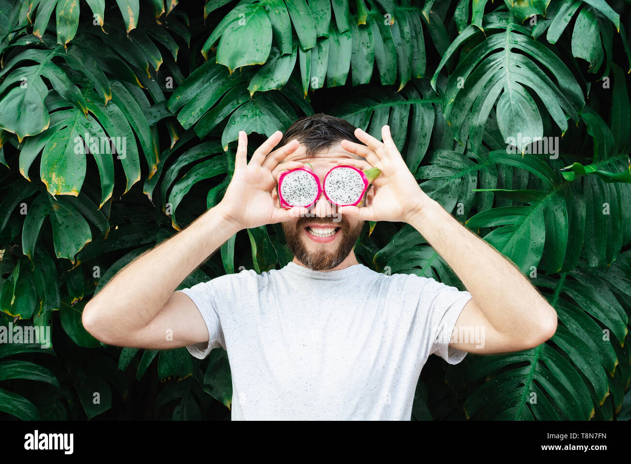 Giovane uomo barbuto holding fette di Pitaya dragon frutto di fronte ai suoi occhi, ridendo. Foglie tropicali sfondo, copia dello spazio. Foto Stock