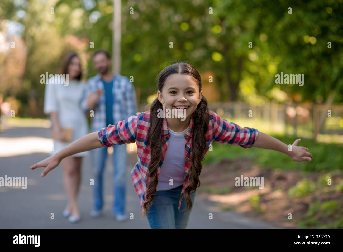 Sorridente Bambina nel parco con le braccia aperte. I genitori sono lo sfondo. Foto Stock