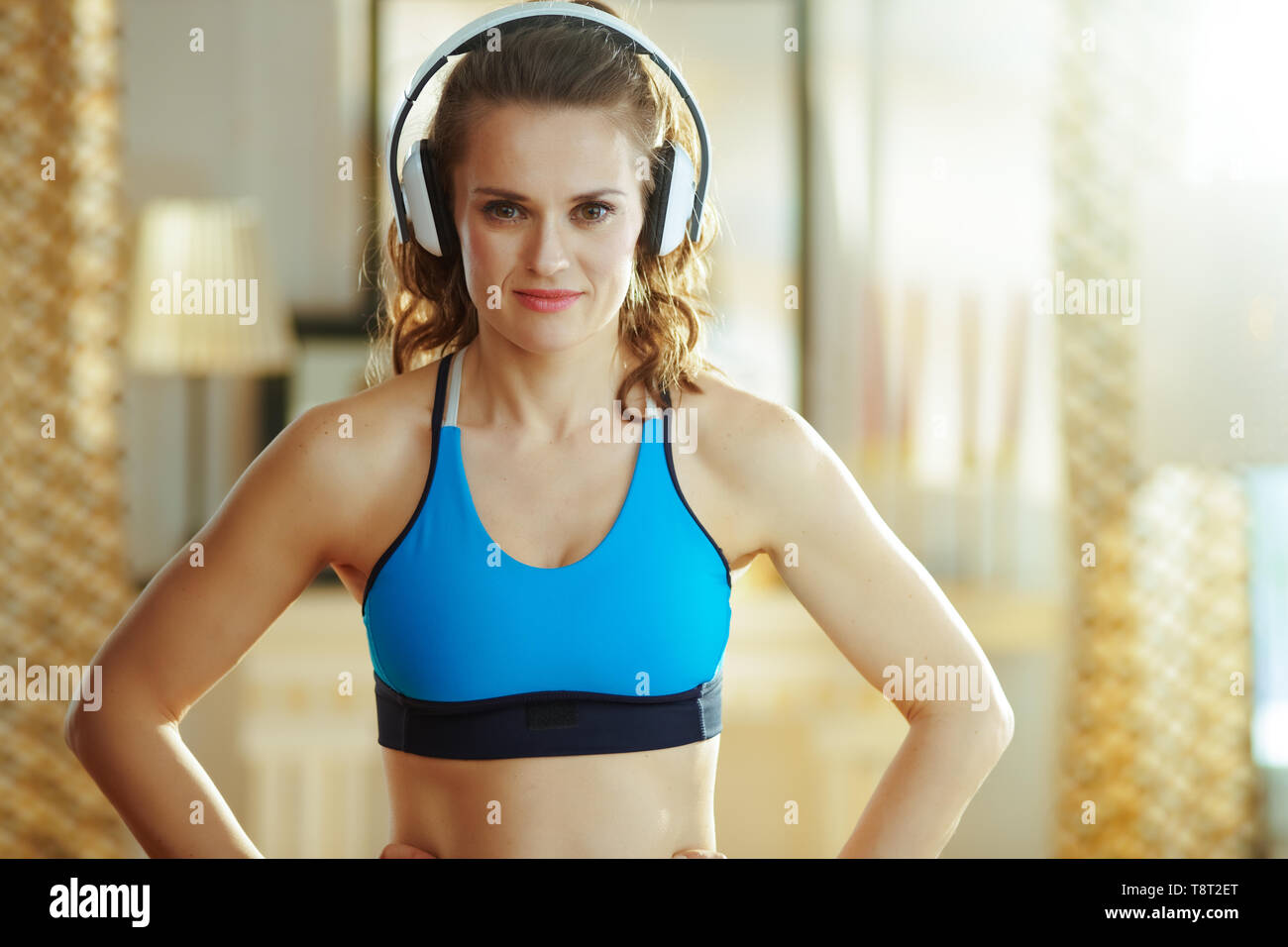 Montare la donna in abiti fitness ascoltando la musica con le cuffie nel soggiorno moderno. Foto Stock