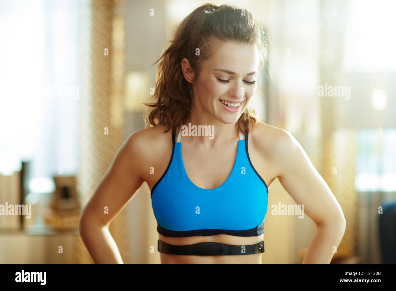 Montare sorridente sport donna in abiti di fitness con monitor per la frequenza cardiaca nel soggiorno moderno. Foto Stock