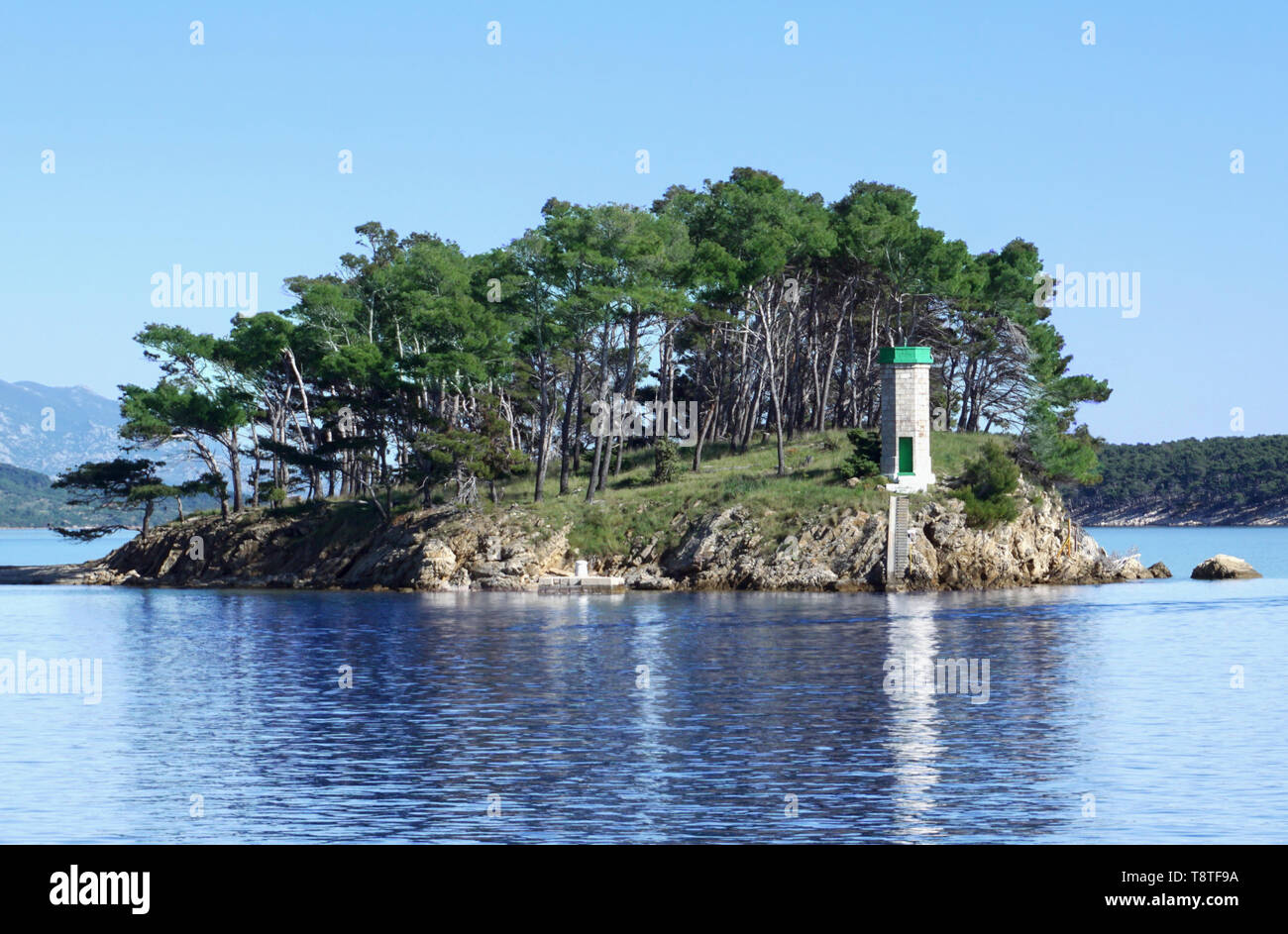 La riflessione di piccolo faro nel bel mare blu superficie nella parte anteriore del porto della città di Rab sul litorale croato, circondato da antichi verde pino Foto Stock