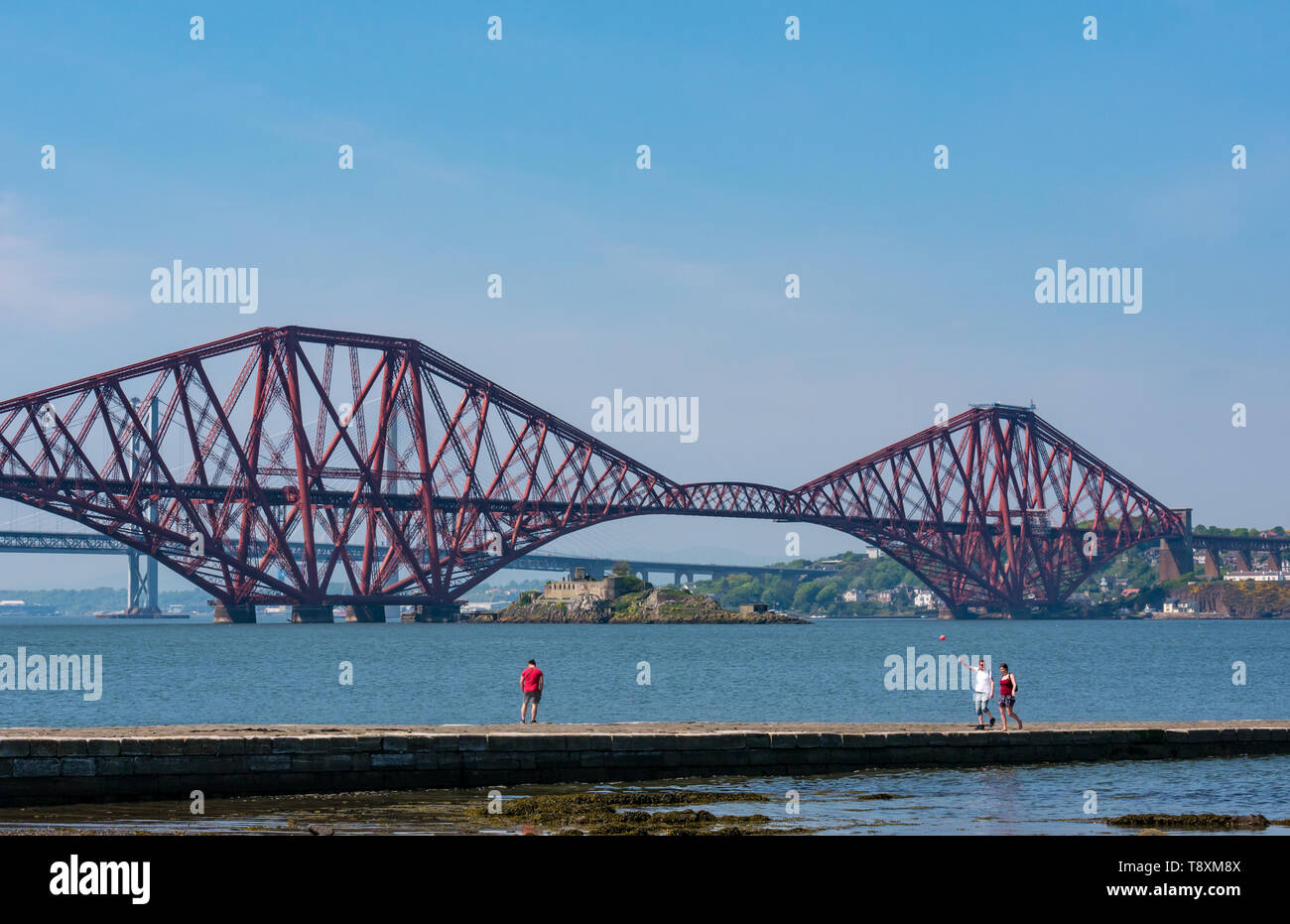 South Queensferry, Scotland, Regno Unito, 15 maggio 2019. Regno Unito: Meteo persone passeggiare lungo il molo in una giornata di sole sul Firth of Forth coast presso il celeberrimo Ponte di Forth Rail e Ponte di Forth Road a distanza Foto Stock
