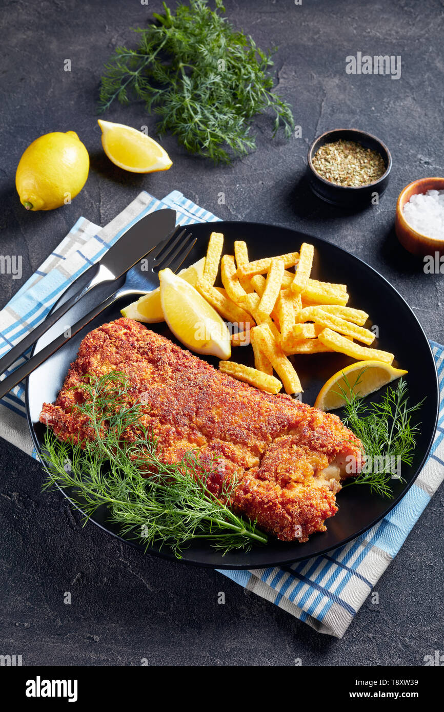Close-up di nasello panato filetto servito con chips, aneto fresco e le fette di limone su una piastra nera su una tavola di cemento, verticale vista da sopra Foto Stock