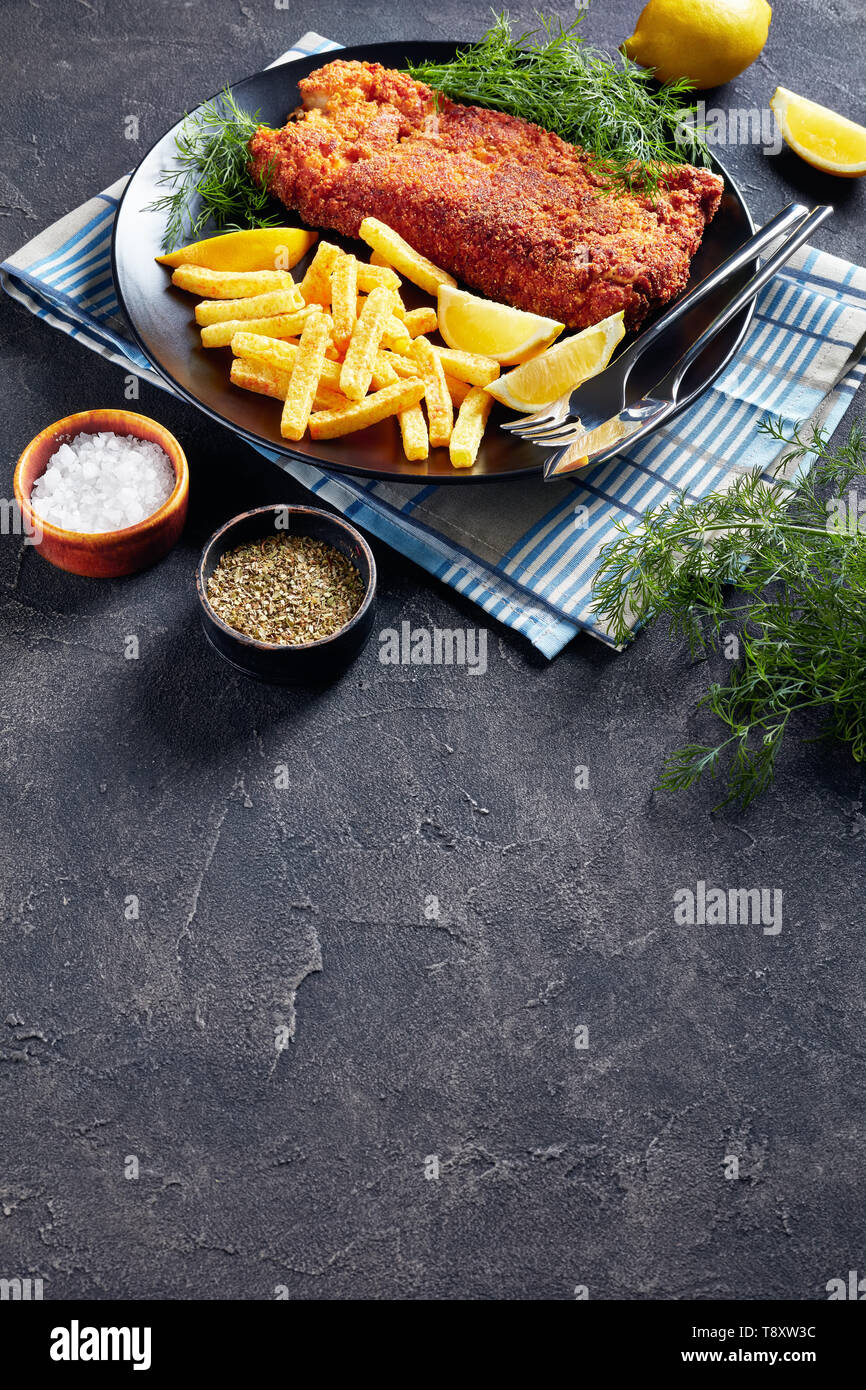 Nasello panato filetto servito con chips, aneto fresco e le fette di limone su una piastra nera su una tavola di cemento, vista verticale dal di sopra, spazio vuoto Foto Stock