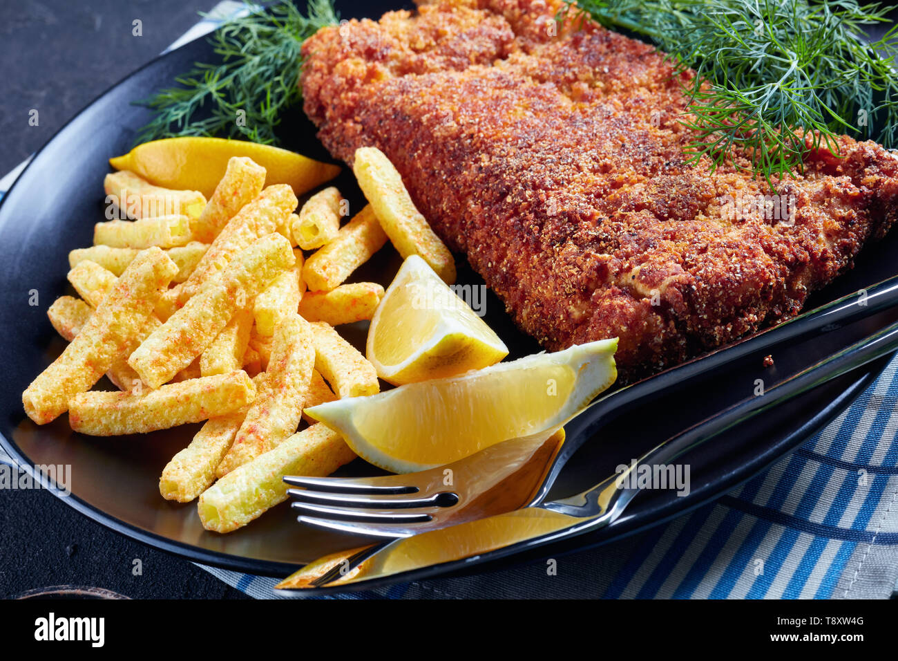 Close-up di nasello panato filetto servito con chips, aneto fresco e le fette di limone su una piastra nera su una tavola di cemento, vista dall'alto Foto Stock