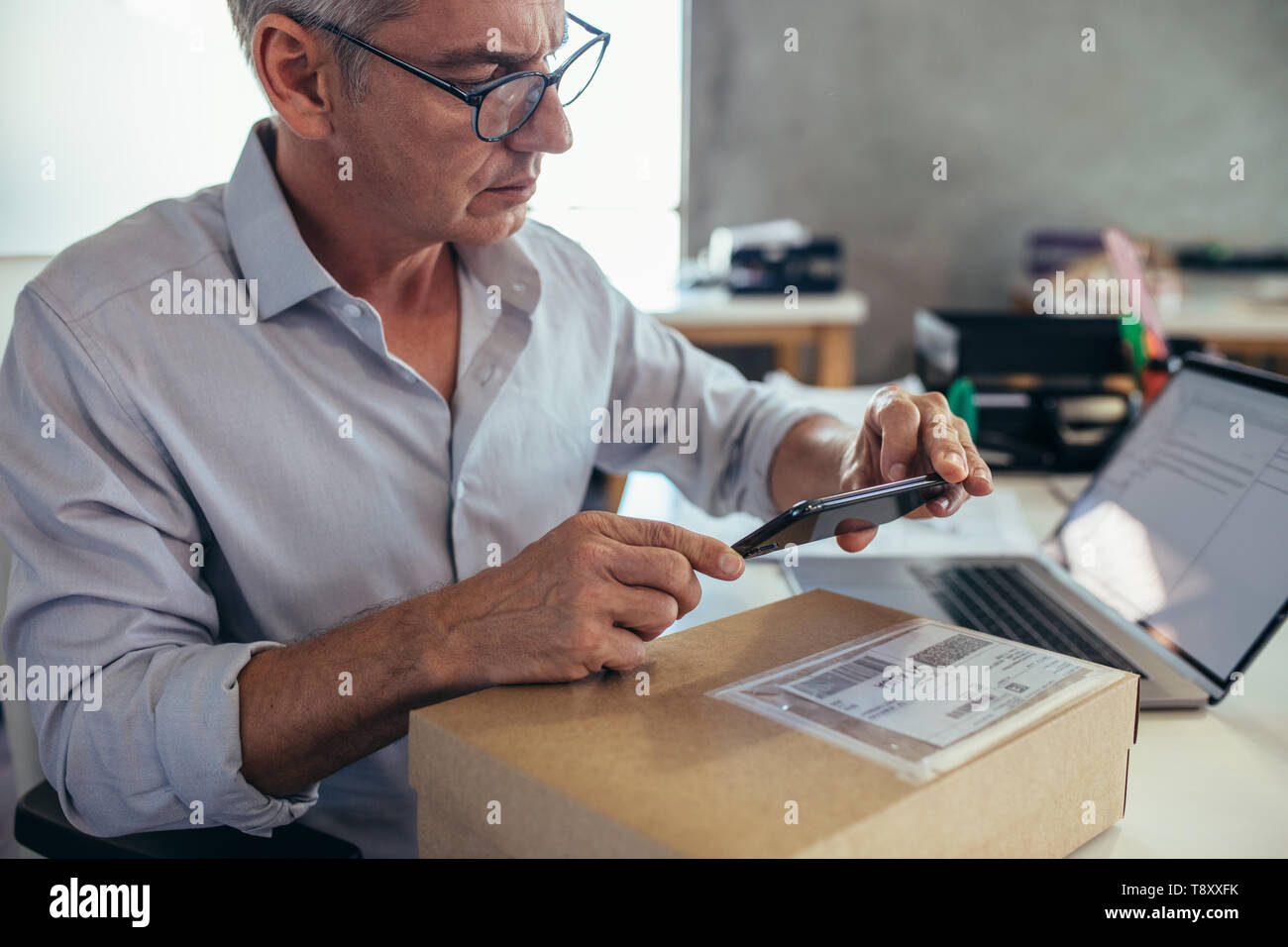 Imprenditore maturo seduto alla sua scrivania e la scansione di una scatola di consegna con il suo telefono cellulare. L'uomo della vendita di prodotti online, la scansione del codice a barre con il suo m Foto Stock