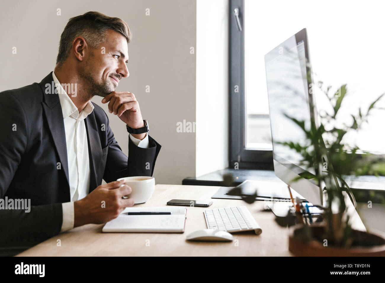 Immagine dell'imprenditore unshaved 30s indossare tuta di bere il caffè mentre si lavora sul computer in ufficio Foto Stock