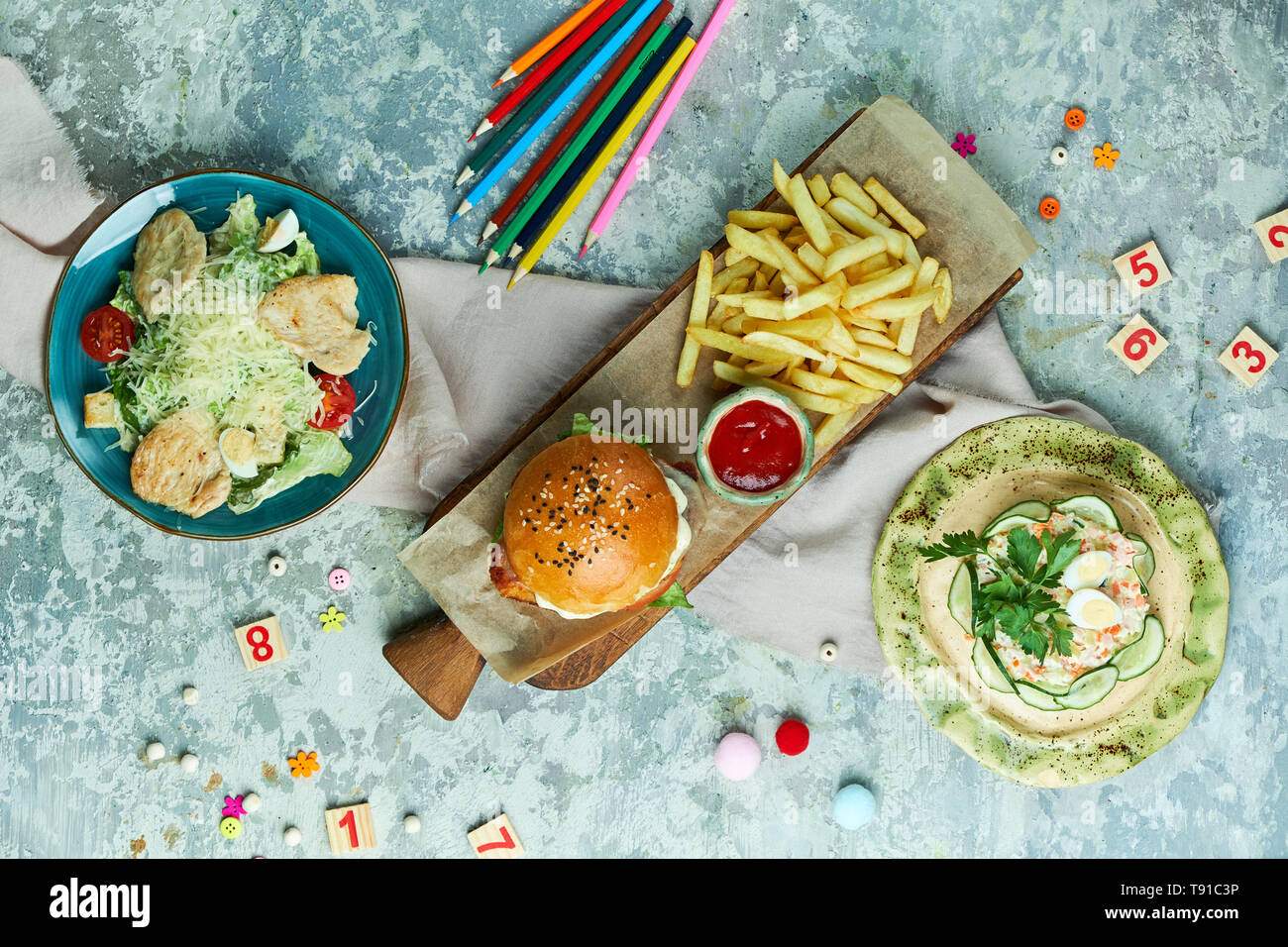 Su un grigio sfondo a trama è una piastra blu con insalata tseat, una tavola di legno con un hamburger e patatine, una piastra di colore giallo con insalata di granchio. Bellissimi piatti di servizio. Menu per bambini nel ristorante. Vista da sopra Foto Stock