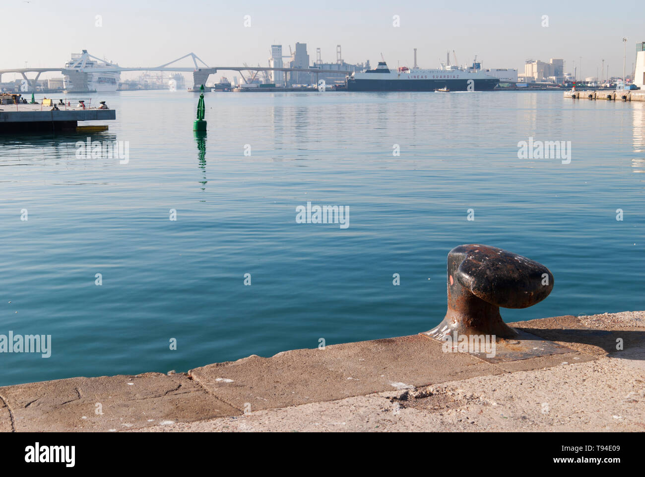 Acque calme all'interno del porto di Barcellona, con un bollard di ormeggio, una boa lontana e il ponte porta d'Europa sullo sfondo. Foto Stock