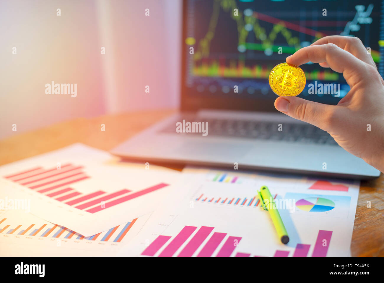 Mano bitcoin di contenimento nella parte anteriore del computer portatile e diagrammi sulla scrivania Foto Stock