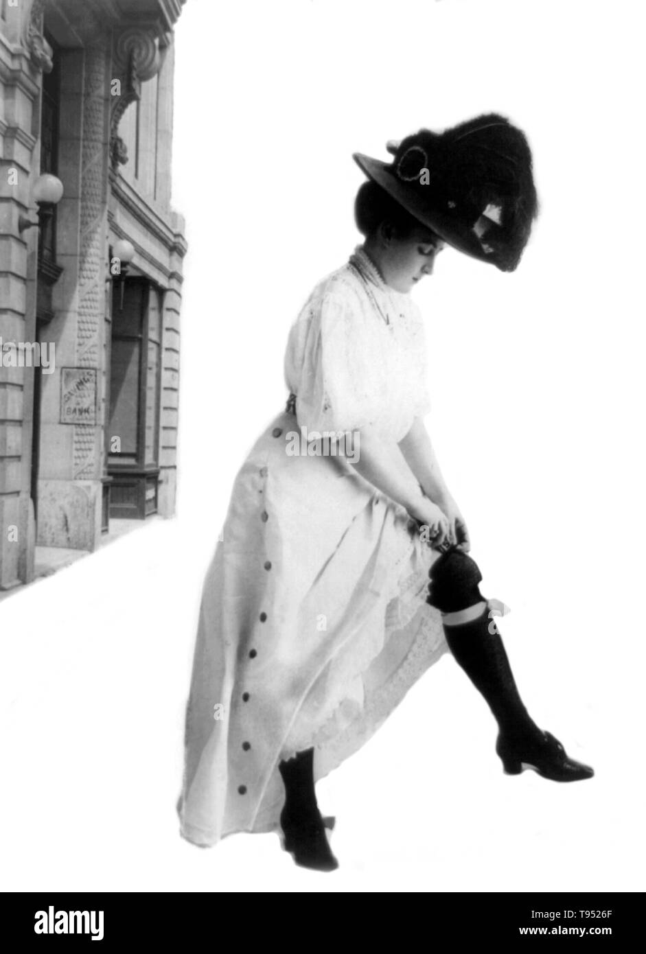 Dal titolo: 'Bonifico Bancario' donna in piedi al di fuori del risparmio bancario; indossando abiti lunghi e lungo cappello piumato; Lei alza il mantello per inserire $5 bill all'interno di lei gartered calza. Fotografata da arte consolidato Company, 1908. Foto Stock