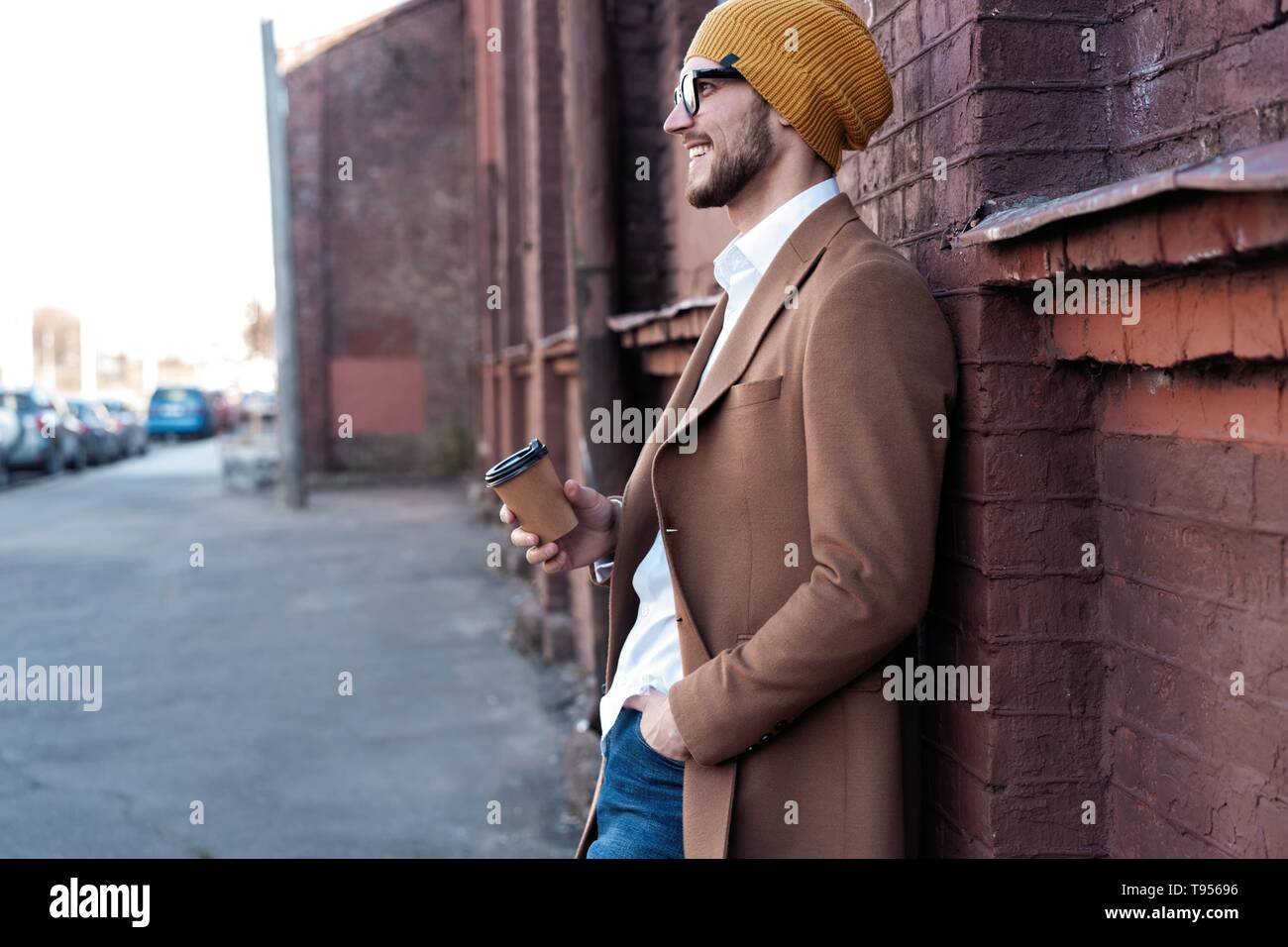 Bel giovane in abbigliamento casual holding tazza monouso e sorridenti mentre si cammina attraverso la strada della citta'. Foto Stock