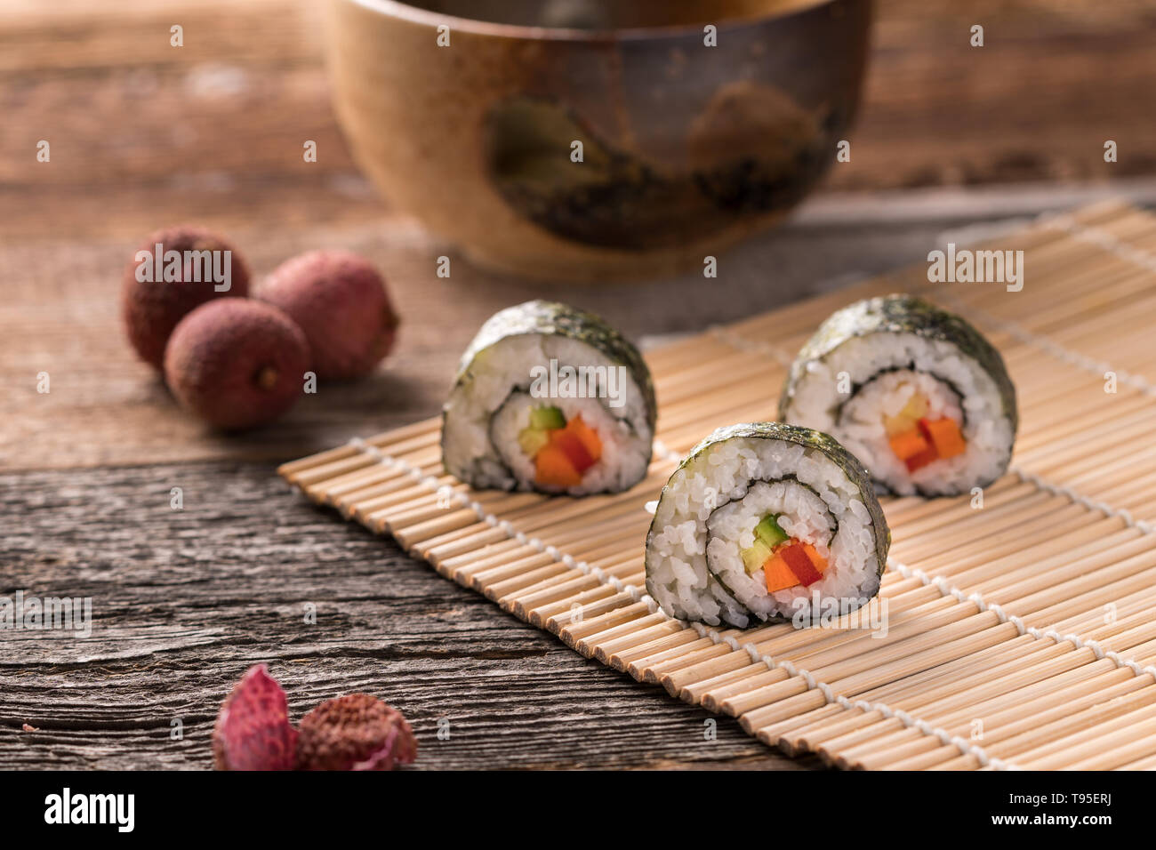 Tre maki sushi rotoli sul tappetino di bambù su una superficie di legno, tradizionali asiatici tee ciotola, litchi e litchi buccia - angolo di visualizzazione, sfondo sfocato, landscap Foto Stock
