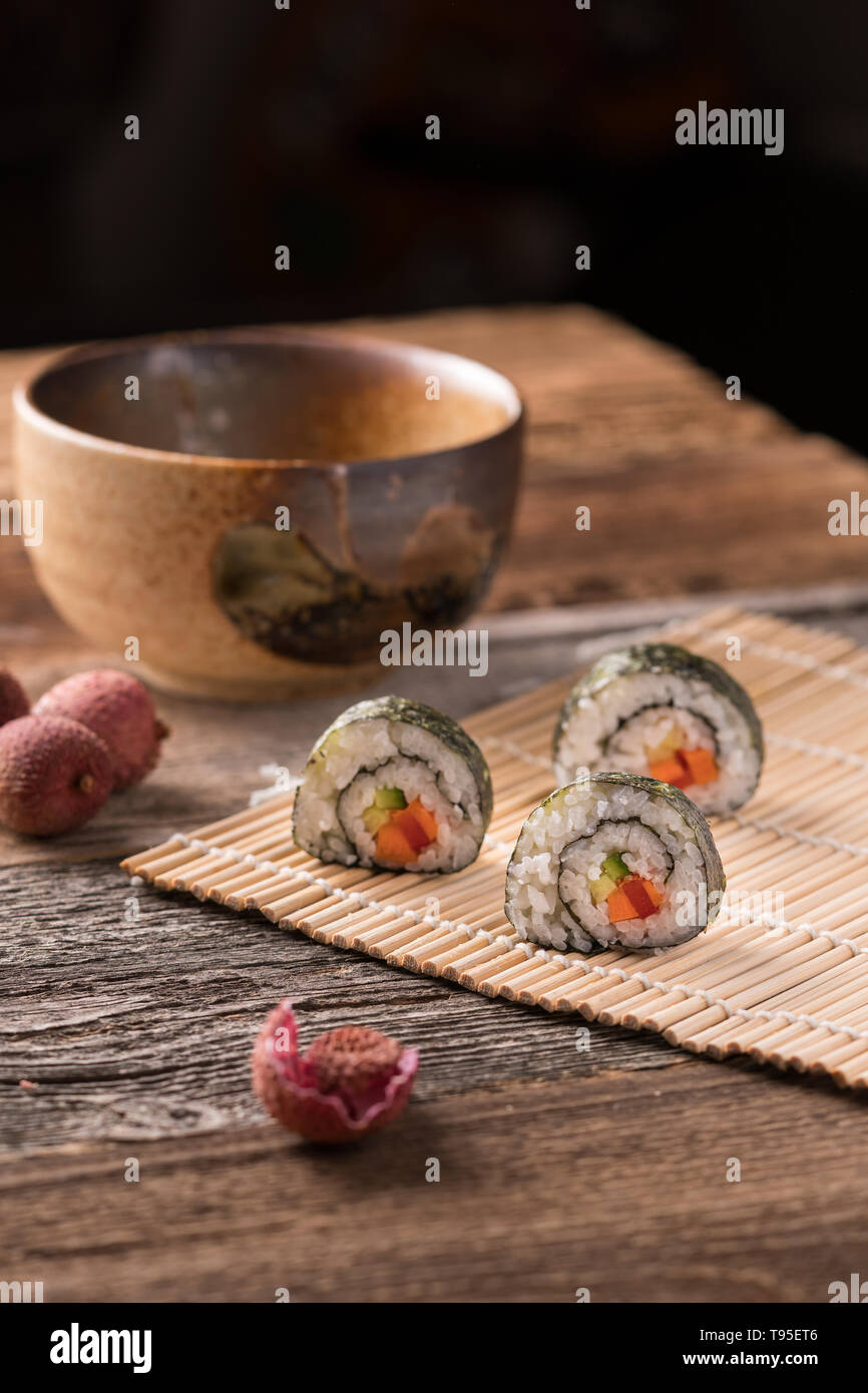 Tre maki sushi rotoli sul tappetino di bambù sul tavolo di legno, tradizionali asiatici tee ciotola, litchi e litchi buccia - angolo di visualizzazione, sfondo sfocato, verticale f Foto Stock