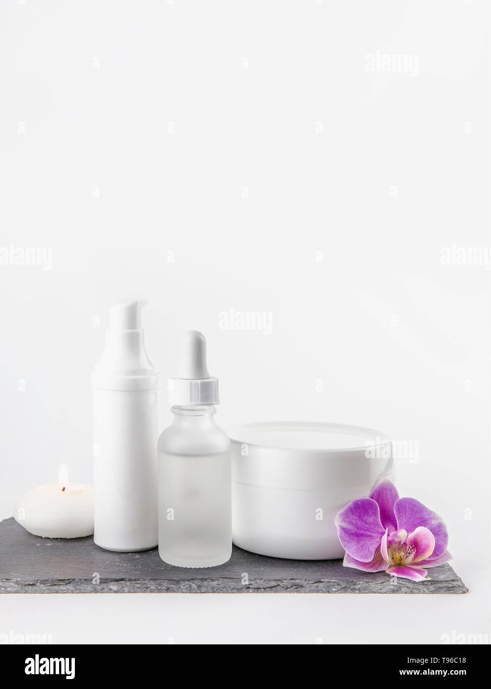 Materiale di diversa forma e dimensione prodotto cosmetico crema, contenitori di olio su sfondo bianco in piedi sulla pietra nera base, rosa fioritura di orchidee. Foto Stock