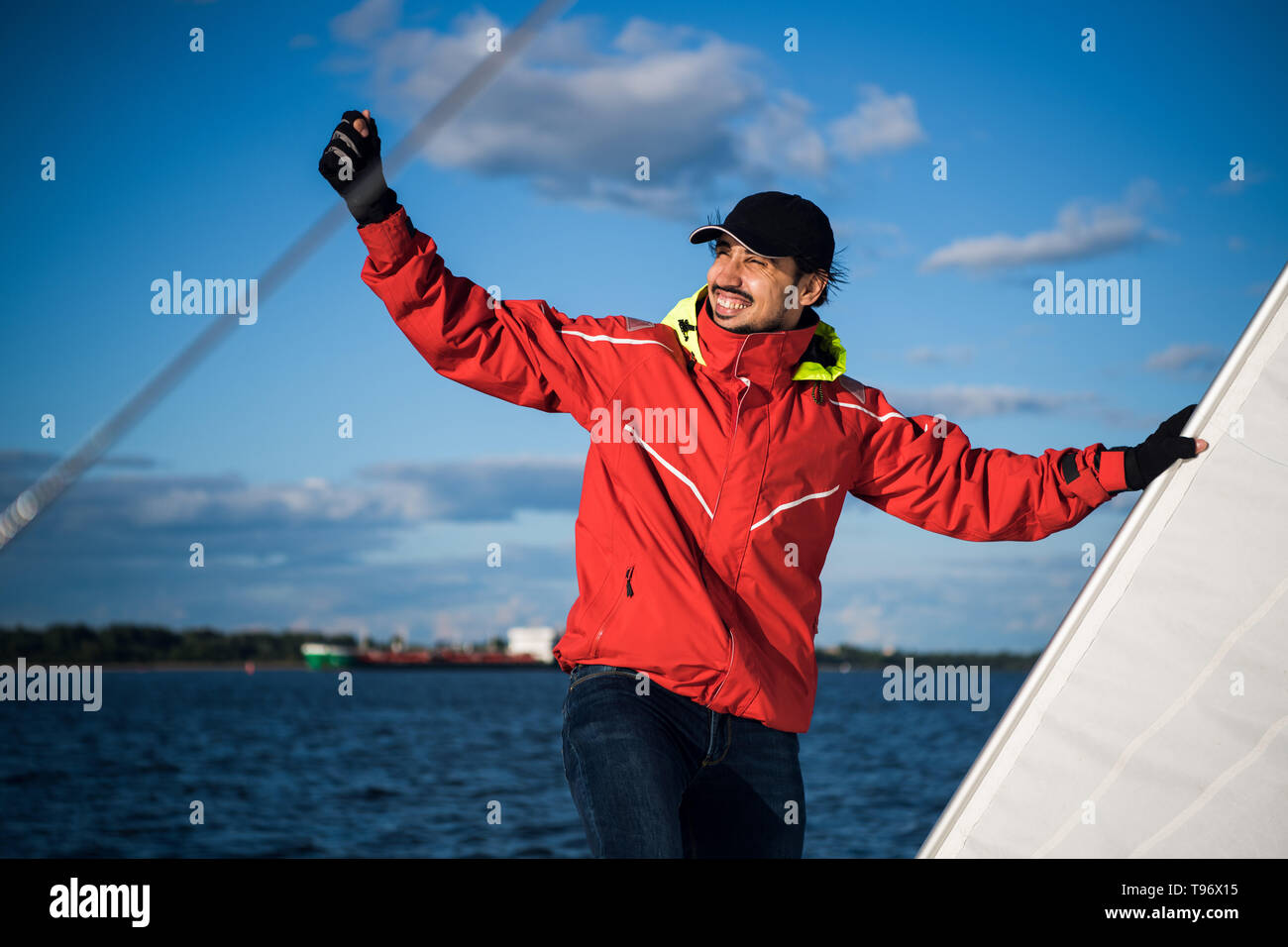 Giovane uomo skipper in rosso giacca a vento e il cappuccio nero imposta le vele su uno yacht a vela. Foto Stock
