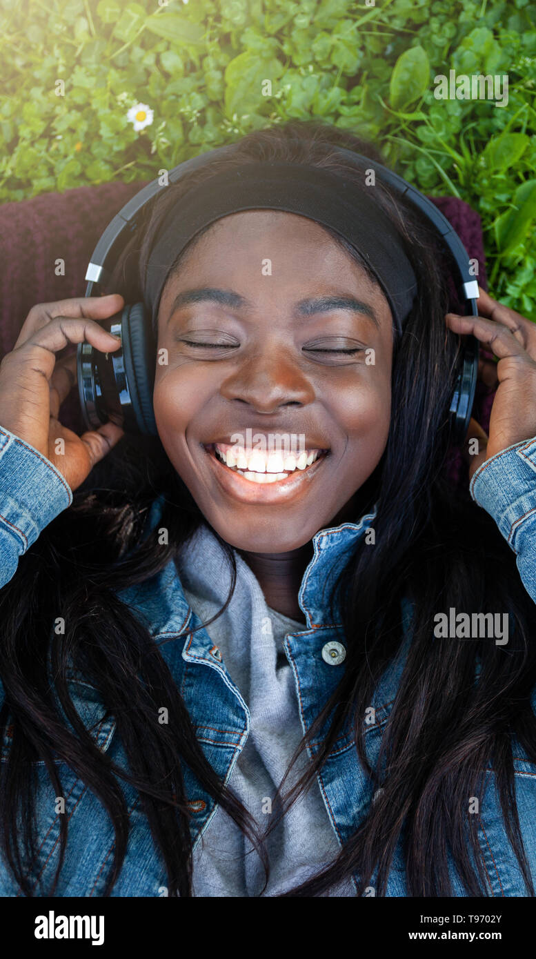 Giovane donna africana di ridere mentre si ascolta la musica con le cuffie giacente su erba verde all'esterno. Foto Stock