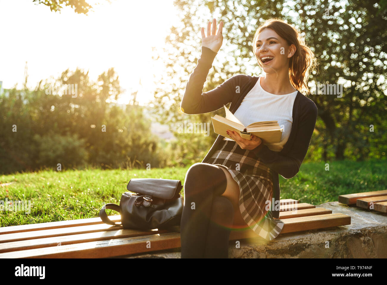 Bella ragazza adolescente indossano uniformi scolastiche libro lettura mentre è seduto su una panchina nel parco, agitando la mano Foto Stock