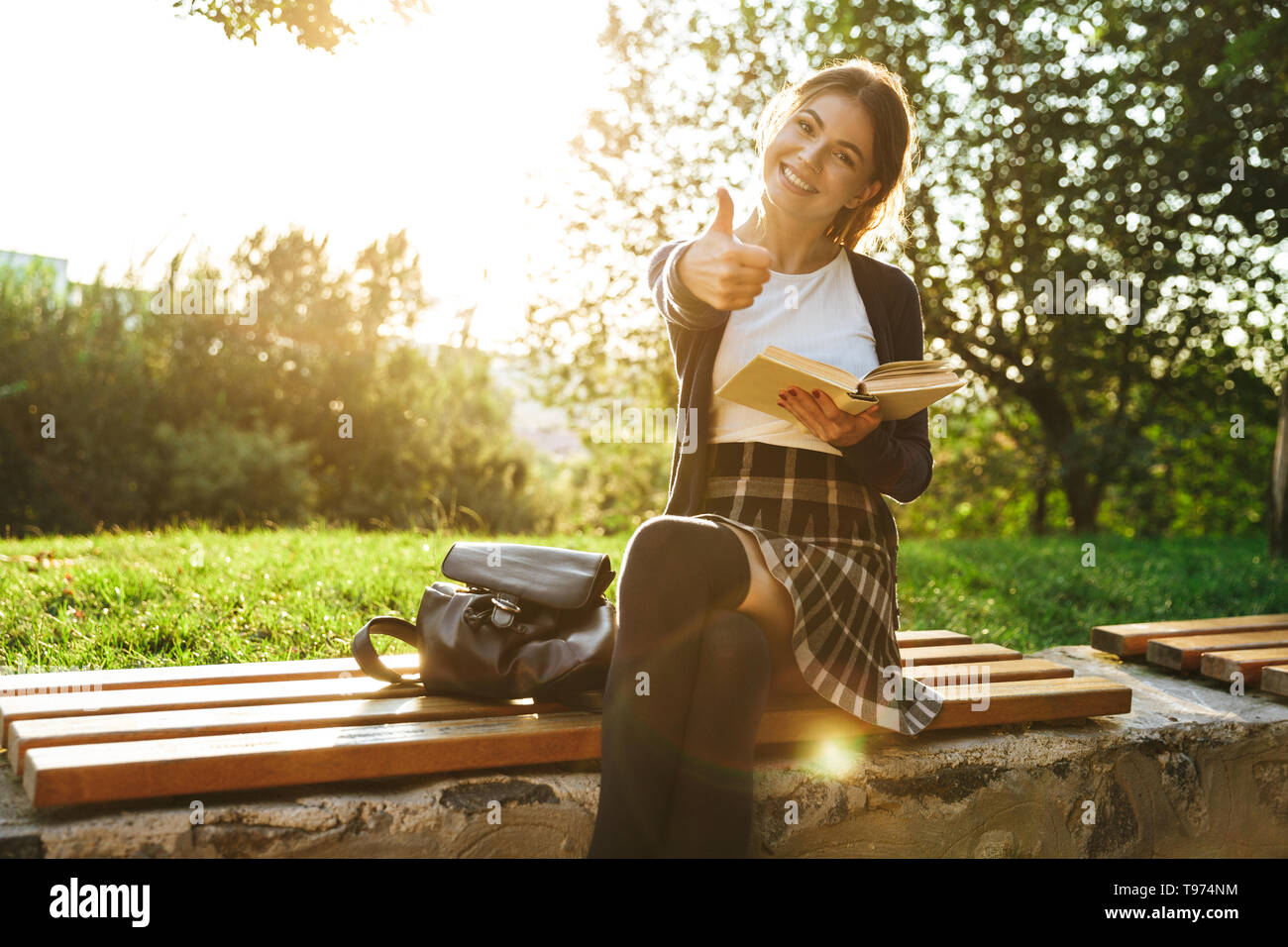 Bella ragazza adolescente indossano uniformi scolastiche libro lettura mentre è seduto su una panchina nel parco, pollice in alto Foto Stock