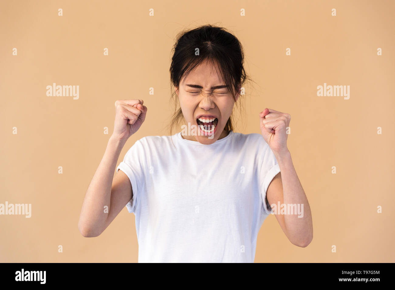Foto di emozionale coreano donna che indossa base t-shirt sorridente e stringendo i pugni isolate su sfondo beige in studio Foto Stock