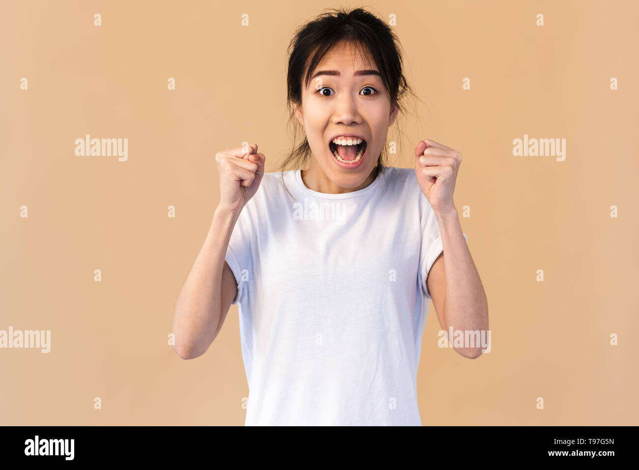 Foto di lieti coreano donna che indossa base t-shirt sorridente e stringendo i pugni isolate su sfondo beige in studio Foto Stock