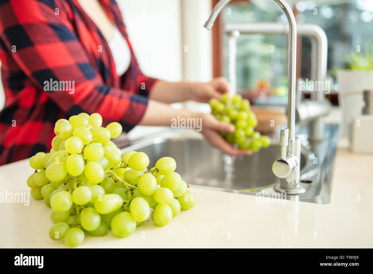 Uva verde sul primo piano e una donna sciacqua l'uva verde con acqua in cucina. Foto Stock