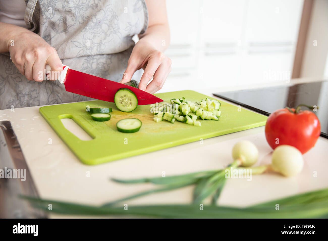Donna in Grembiule da cucina taglia il cetriolo sul tagliere con coltello rosso con pomodoro ed erba cipollina sul primo piano. Foto Stock
