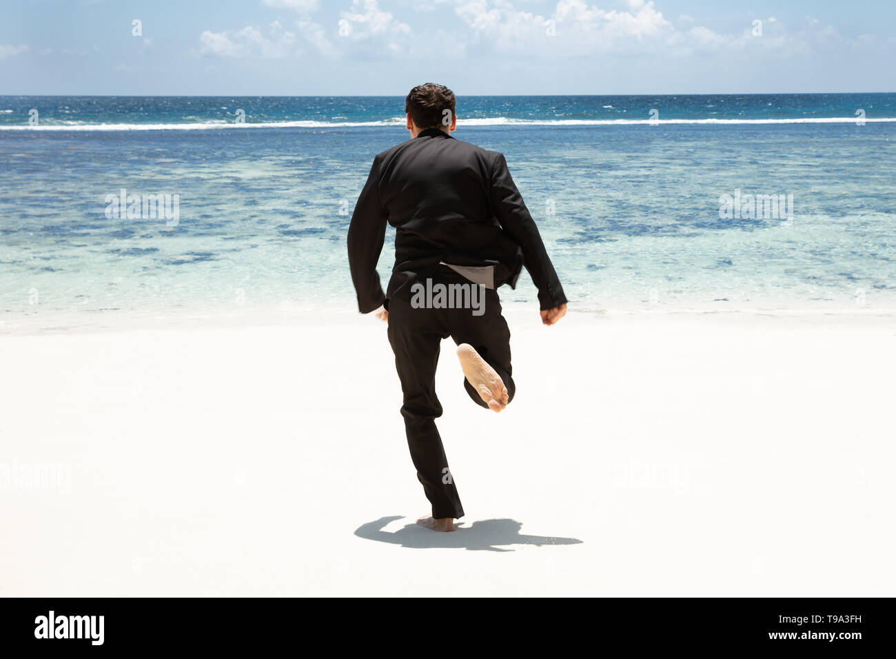 Vista posteriore di un uomo in abito nero correre a piedi nudi verso il mare in spiaggia Foto Stock