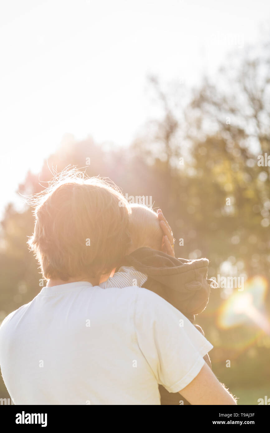 Padre cullano il suo bambino tra le sue braccia nel caldo bagliore del sole come si cancella le cime degli alberi circostanti. Foto Stock