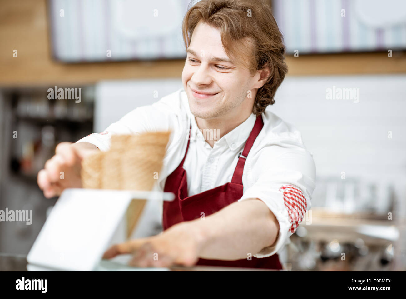 Bello sorridente venditore tenendo cono di cialda durante la preparazione del gelato per il client nella moderna pasticceria Foto Stock