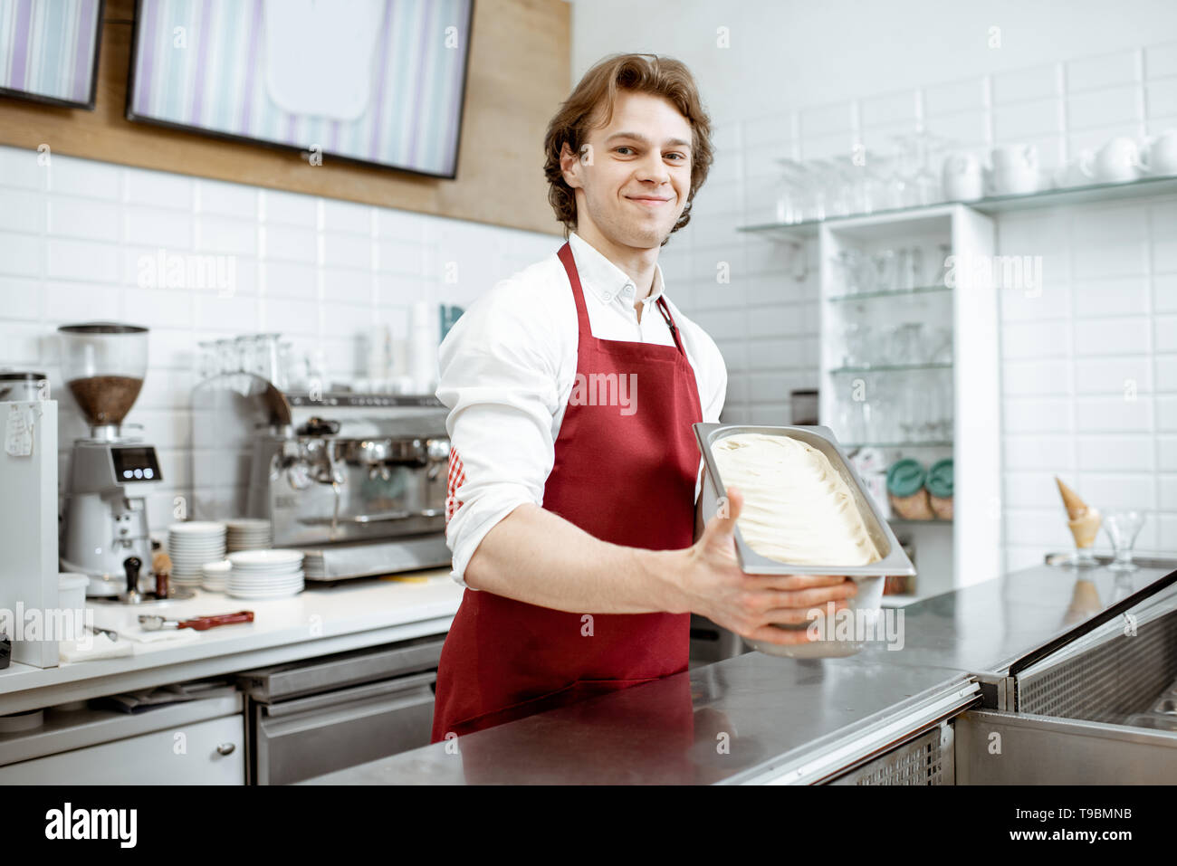 Ritratto di un bel venditore o pasticceria a grembiule rosso tenendo il vassoio con gelato nel negozio Foto Stock