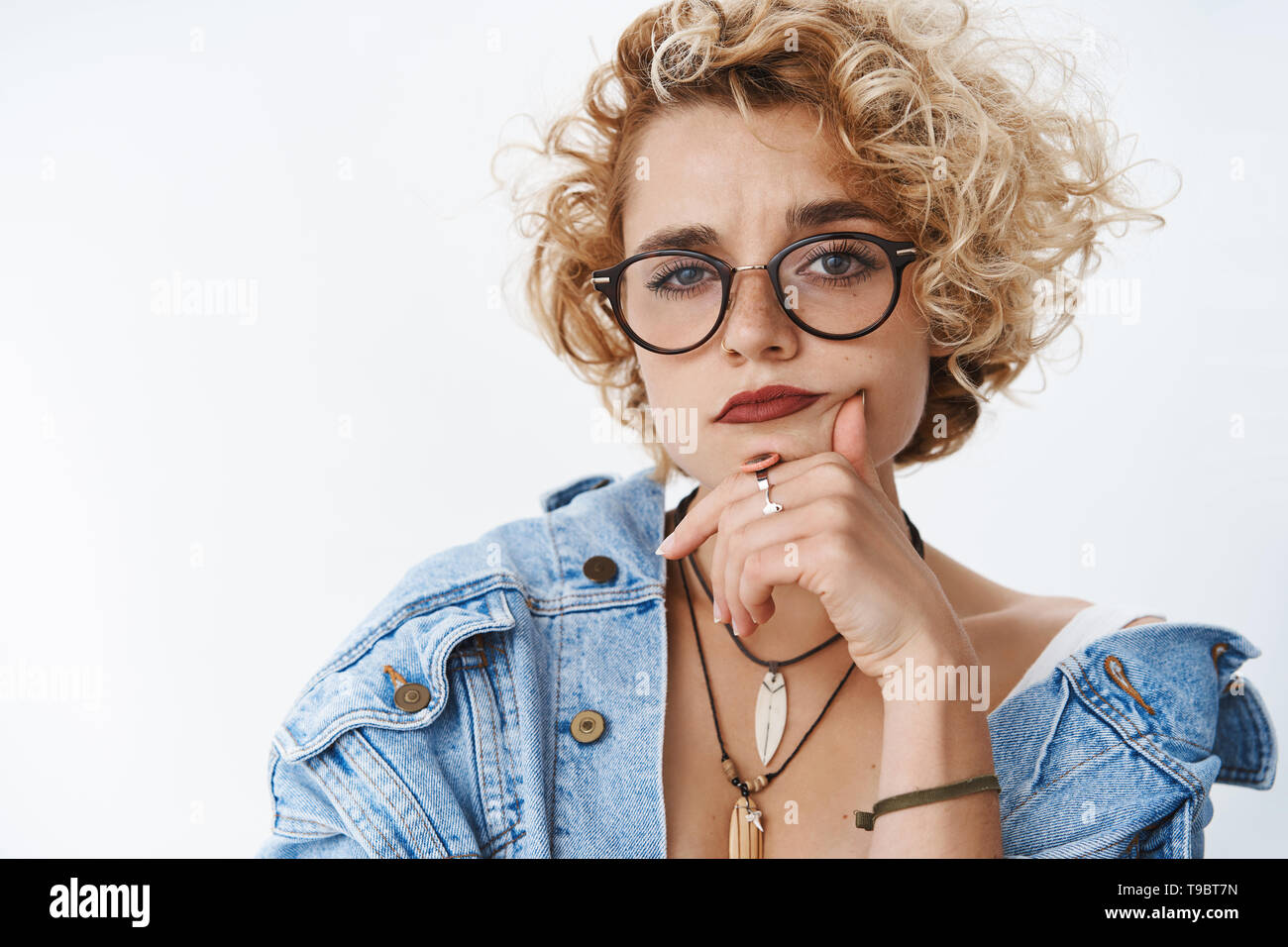 Interrogato ragazza cercando in dubbio e sospetto. Ritratto di incerta attraente ragazza in bicchieri e camicia denim tenendo la mano sul mento Foto Stock