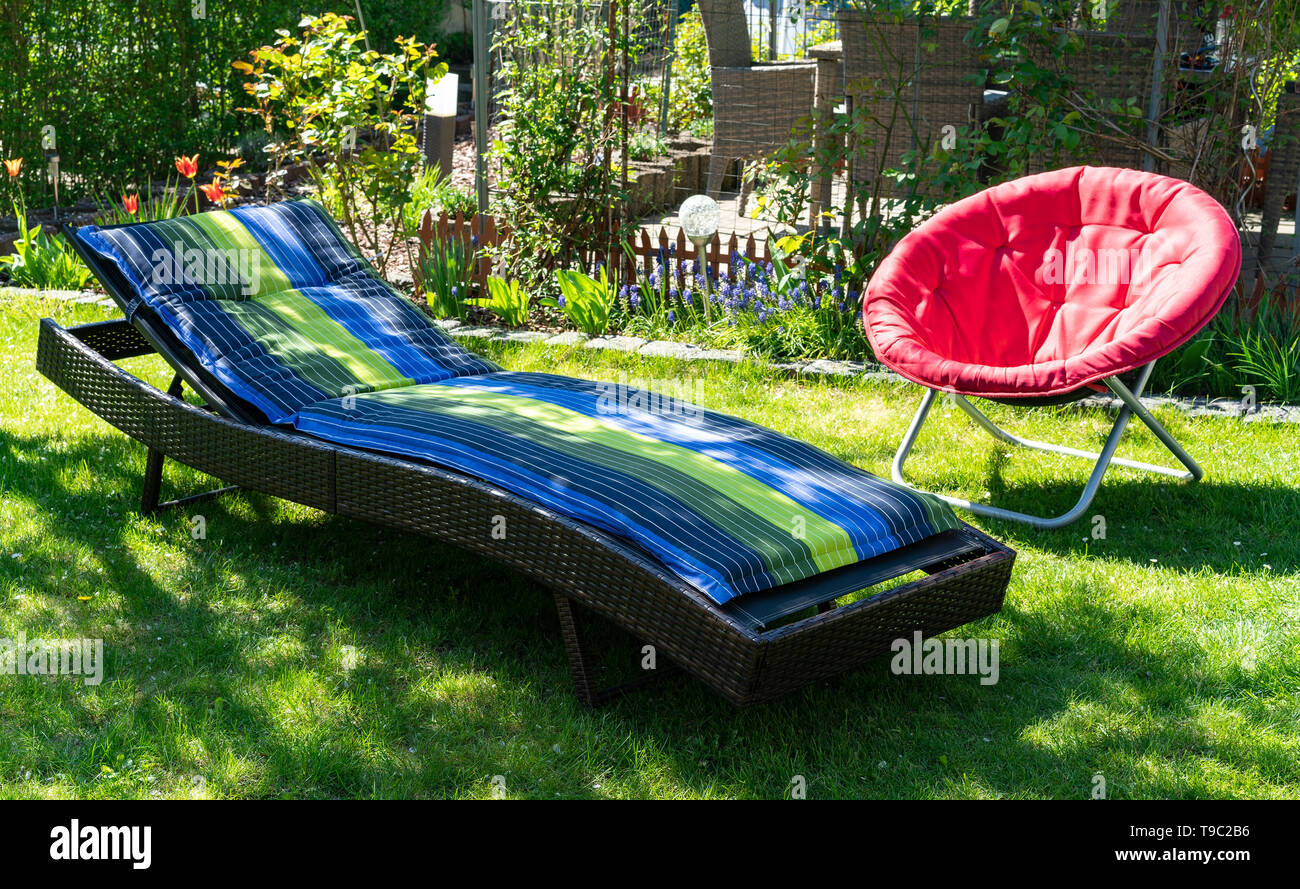 Un blu e verde di lettini e un rosso rotondo rosso luna pieghevole sedia in un giardino verde in piedi nel sole durante la primavera tempo.. Foto Stock