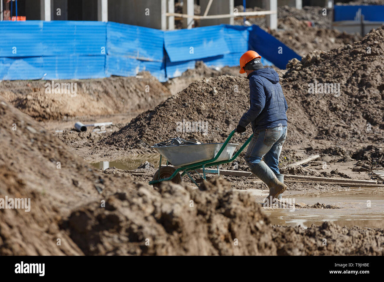 Un lavoratore in un casco porta una carriola contro lo sfondo di detriti di costruzione. Foto Stock