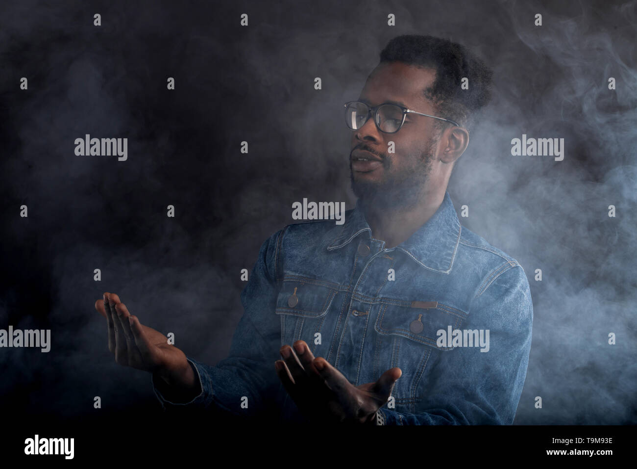Che cos'è. Close up ritratto di sorpreso bello african american uomo in bicchieri, vestito casualmente in piedi nella nube di fumo bianco con i palmi rivolti verso l'alto, Foto Stock