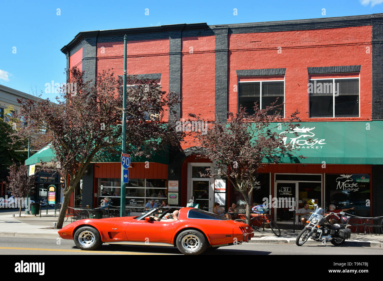 Un uomo causcasion in un rosso Corvette Convertibile unità nel centro di Coeur d'Alene, ID, accanto al mercato delle alci e lounge in vintage rosso e nero b Foto Stock