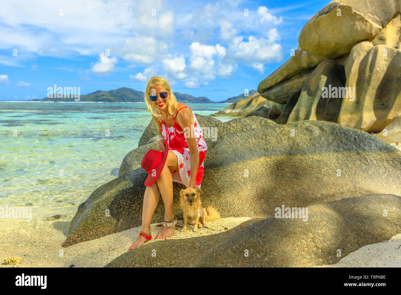 Stile di vita sorridente donna seduta su un massi, tocca un simpatico cane ad Anse Source d'Argent una delle più belle spiagge di La Digue, Seicelle Foto Stock