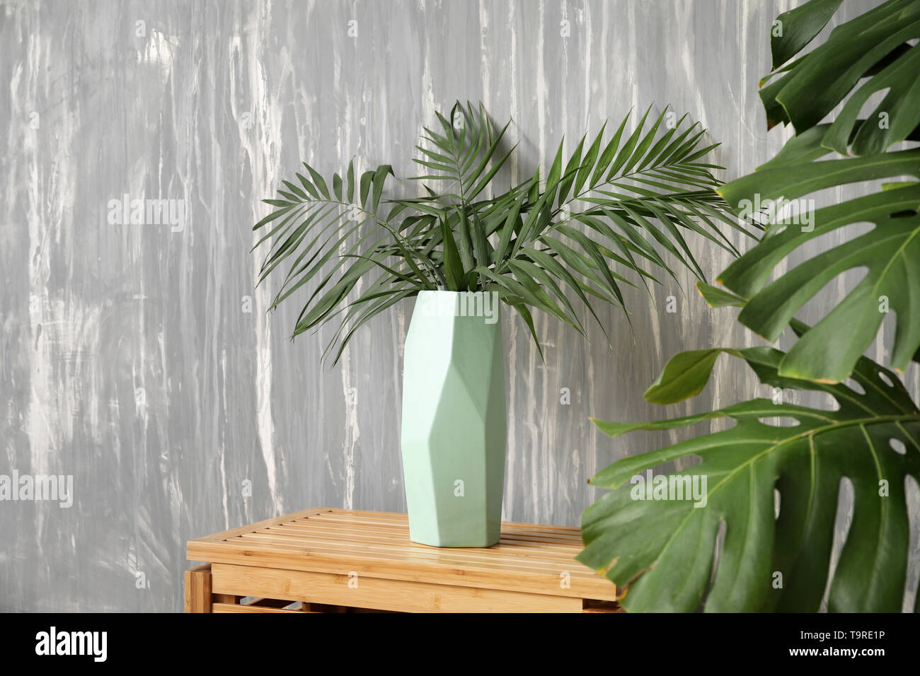 Vaso con verde tropicale, lascia sul tavolo vicino al muro grigio Foto Stock