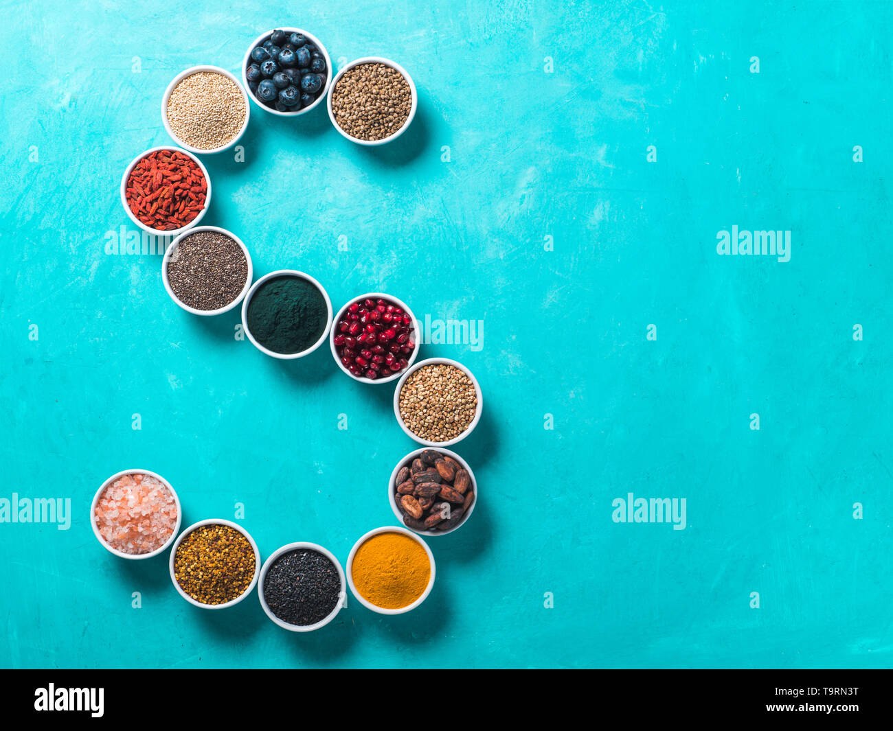 Al di sopra di vista di vari superfoods in piccolo vaso in forma S lettera su sfondo blu. Super come chia,spirulina,fava di cacao,goji, canapa, mirtillo, quinoa, polline di api,sesamo nero,curcuma. Vista superiore Foto Stock