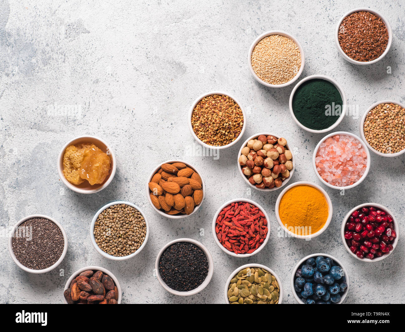 Varie superfoods nella piccola ciotola di cemento grigio Sfondo. Super come chia, spirulina, materie fava di cacao, goji, canapa, quinoa, polline di api, sesamo nero, curcuma. Copia spazio per il testo. Vista superiore, flat-lay. Foto Stock