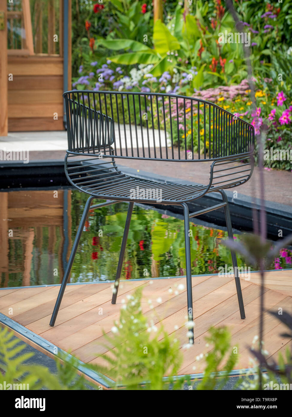 Filo metallico sedia sul giardino decking con stagno, piante e serra in legno in background Foto Stock