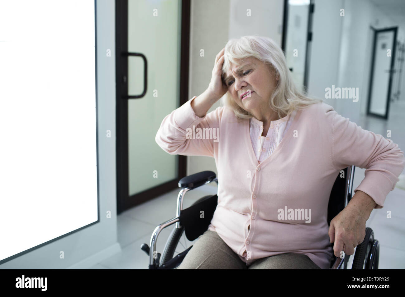 Disperata donna senior tenendo la sua testa seduto nella sedia a rotelle all'ospedale corridoio. La tristezza Foto Stock