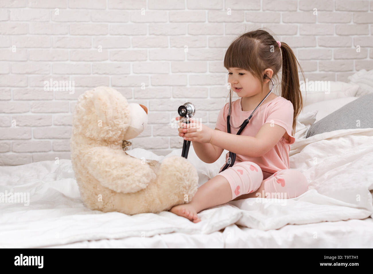 Carina ragazza bambino la riproduzione medico con Teddy bear a casa. ragazza ascolta uno stetoscopio per giocattolo. Foto Stock