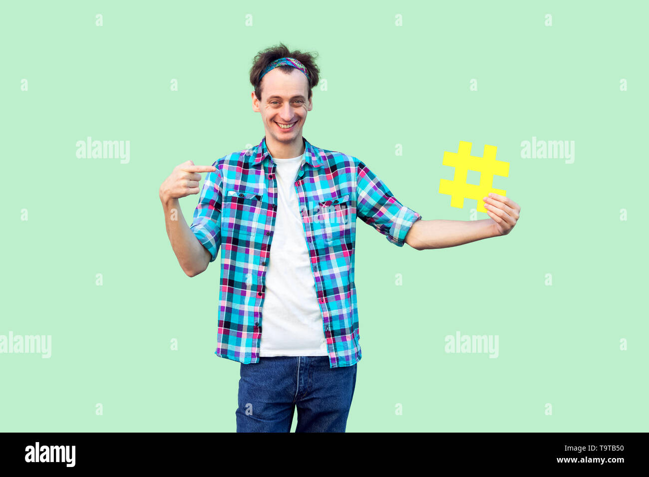 Fate uso di hashtag? Felice positivo Giovane Uomo in camicia a scacchi tenendo una grande giallo tag hash di segno e puntando con il dito su di essa. Indoor, isolare Foto Stock