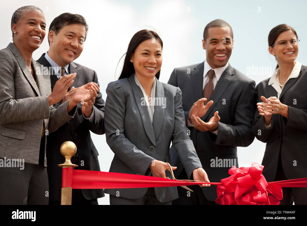 Sorridente di mezza età imprenditrice esegue la cerimonia del taglio del nastro di un nuovo sviluppo come i suoi colleghi guardano su felicemente e applaudire. Foto Stock